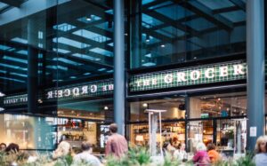 Udsigt til en butik med navnet "The Grocer", moderne glasfacade og indretning, hvor kunder sidder udenfor og nyder stemningen.